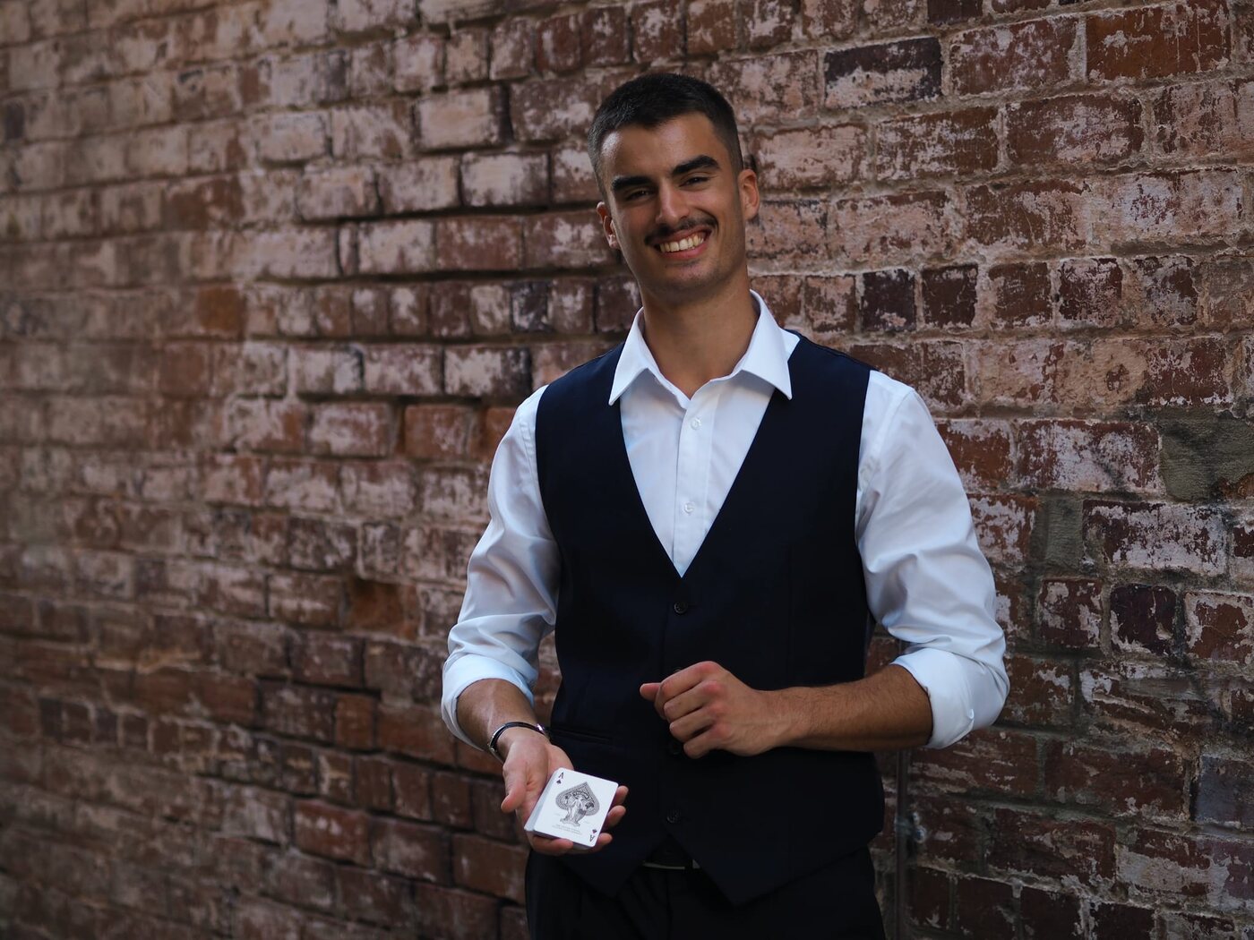 Dominic Miles smiling in a waistcoat, holding the Ace of Spades against a brick wall